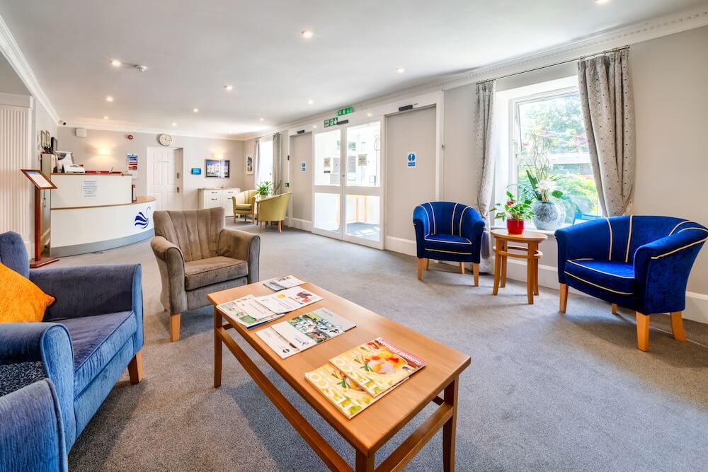 Waiting room with blue, yellow, and beige armchairs, a table with newspapers, and a front desk.