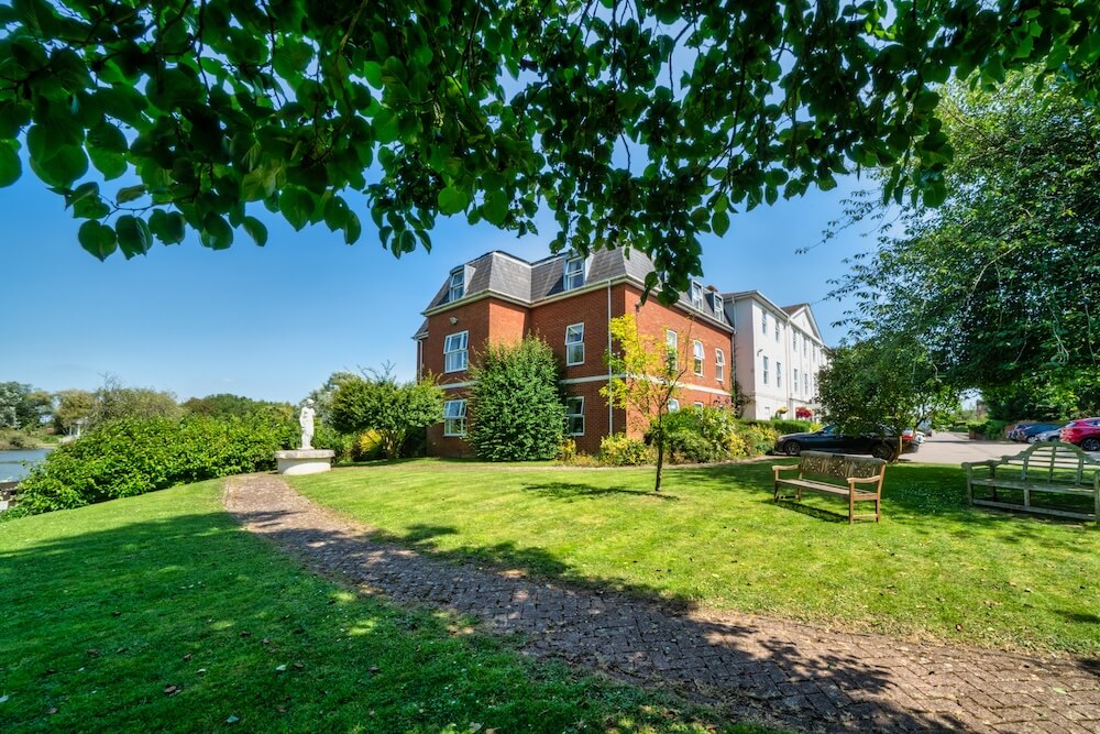 Side view of a care home from the garden, showing a green lawn, bench, tree, and sculpture.