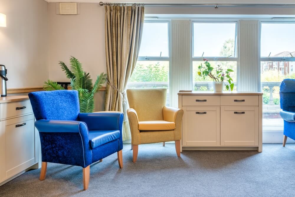 Room with blue and yellow armchairs, a cupboard, and a large window adorned with golden curtains.