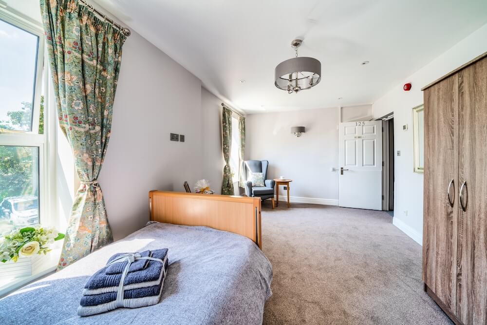 Room view featuring a bed with neatly folded towels on top, a blue armchair with a side table, a wardrobe, and two windows dressed with flowery curtains.